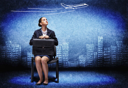 Young upset businesswoman sitting on chair with briefcase in handsの写真素材