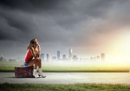 Young pretty girl traveler sitting on suitcase aside of roadの写真素材