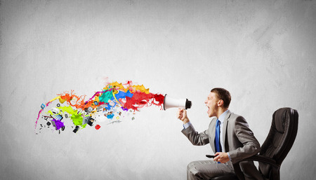 Young angry businessman sitting on chair and screaming in megaphoneの写真素材