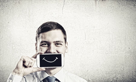 Young handsome businessman hiding mouth behind mobile phoneの写真素材