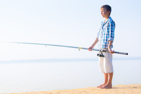 Boy in blue shirt standing on a pier with a fishing rod by the seaの写真素材