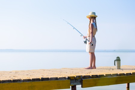 Girl in a dress and a hat with a fishing rod fishing from the pierの写真素材