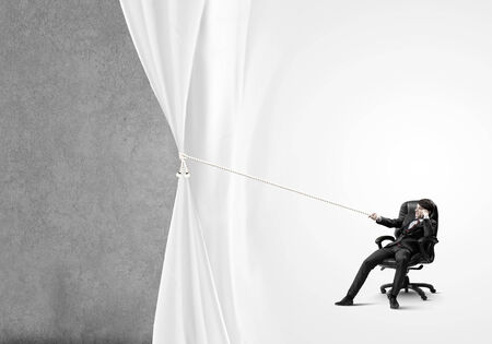 Young businessman sitting in chair and pulling curtain with ropeの写真素材