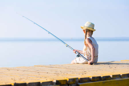 Girl in a dress and a hat with a fishing rod fishing from the pierの写真素材