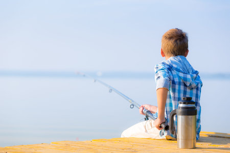 Boy in blue shirt sit on a pier with a fishing rod by the sea
