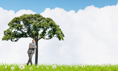 Young relaxed businessman leaning on green treeの写真素材