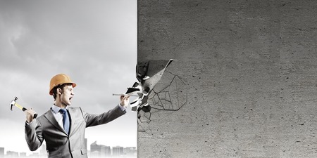 Young businessman in suit and hardhat hammering nail in wallの写真素材