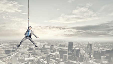 Young businessman hanging on rope above cityの写真素材