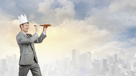 Young handsome businessman wearing white paper crown and looking in spyglassの写真素材