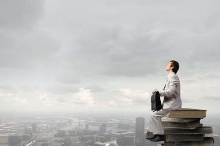 Young businessman with suitcase sitting on pile of old booksの写真素材