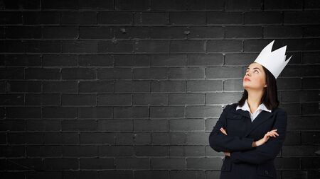 Young businesswoman in paper crown standing with arms crossed on chestの写真素材