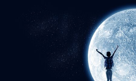 Rear view of young woman with hands up looking at moon in skyの写真素材