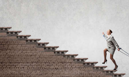 Young businesswoman with ropes on hands climbing ladderの写真素材