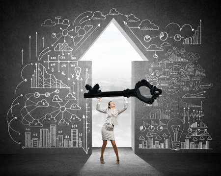 Young businesswoman and keyhole at background lifting big key above headの写真素材