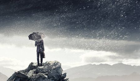 Back view of businessman with umbrella and suitcase standing on mountain topの写真素材