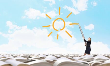 Young businesswoman with big pencil standing on pile of booksの写真素材