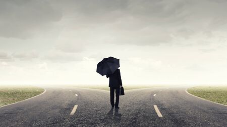 Back view of businessman with umbrella and suitcase walking on roadの写真素材