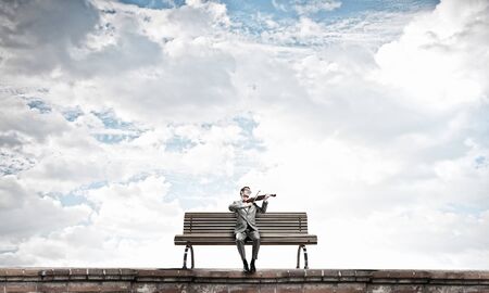 Young man in suit sitting on bench and playing violinの写真素材