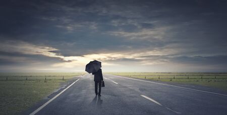 Back view of businessman with umbrella and suitcase walking on roadの写真素材