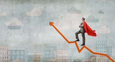 Young man in super hero costume standing on increasing graphの写真素材