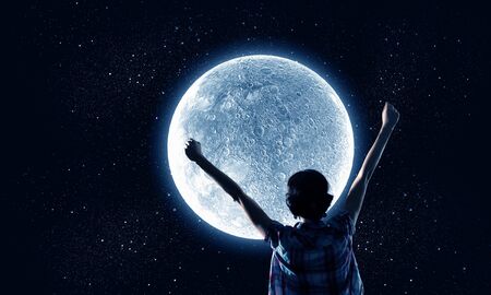 Rear view of young woman with hands up looking at moon in skyの写真素材