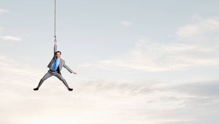 Young businessman hanging on rope high in skyの写真素材