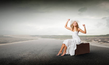 Woman in white long dress and hat sitting on her luggage on asphalt  roadの写真素材