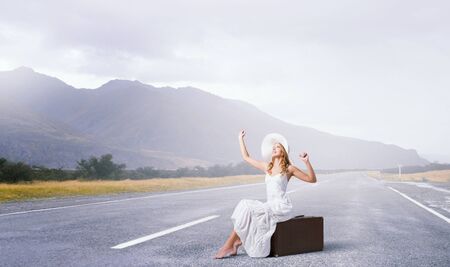 Woman in white long dress and hat sitting on her luggage on asphalt  roadの写真素材