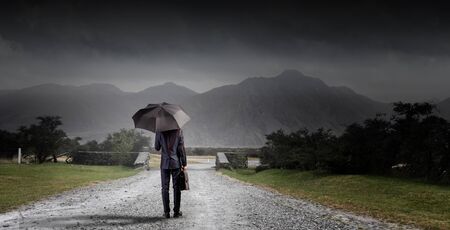 Back view of businessman with umbrella and suitcase walking on roadの写真素材
