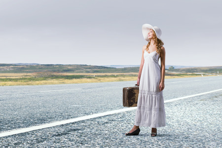 Woman with suitcase in white long dress and hat on asphalt  roadの写真素材