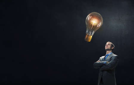 Young confident businessman with arms crossed on chest looking at light bulbの写真素材