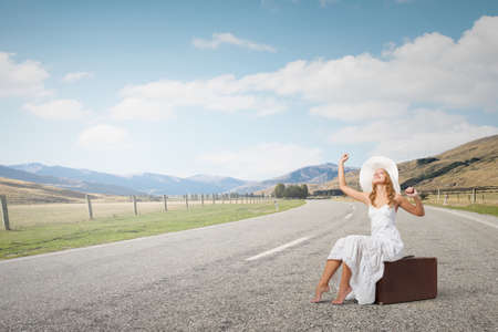 Woman in white long dress and hat sitting on her luggage on asphalt  roadの写真素材