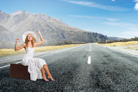 Woman in white long dress and hat sitting on her luggage on asphalt  roadの写真素材