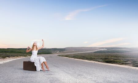 Woman in white long dress and hat sitting on her luggage on asphalt  roadの写真素材