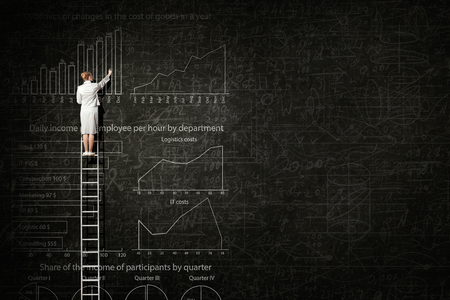 Back view of businesswoman standing on ladder and drawing sketch on wallの写真素材
