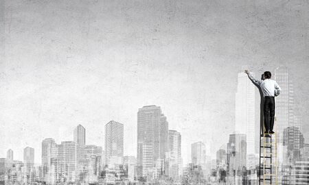 Rear view of businessman standing on ladder and drawing buildingsの写真素材