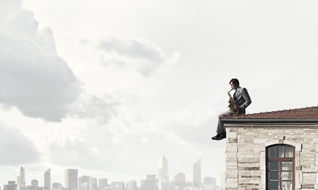 Young man sitting on roof and playing saxophoneの写真素材