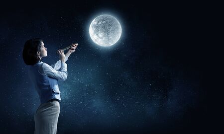 Young businesswoman looking in spyglass on full moon in skyの写真素材