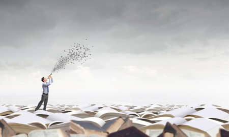 Young carefree businessman on pile of old books playing pipeの写真素材