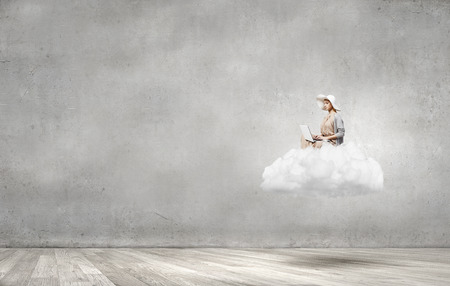 Woman in dress and hat sitting on cloud and working on laptopの写真素材