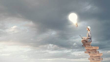 Woman in dress and hat sitting on books and working on laptopの写真素材