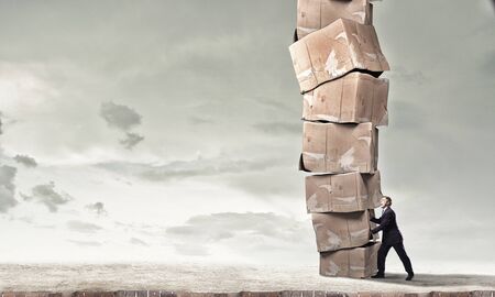 Young businessman making effort to move stack of carton boxes