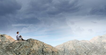 Young thoughtful businessman sitting alone on rock topの写真素材