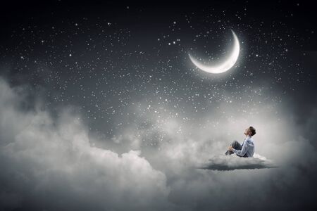 Businessman in suit sitting on cloud in isolation and looking at moonの写真素材