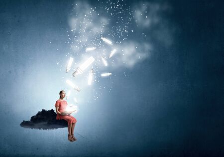 Young woman in dress with old book in hand sitting on cloudの写真素材