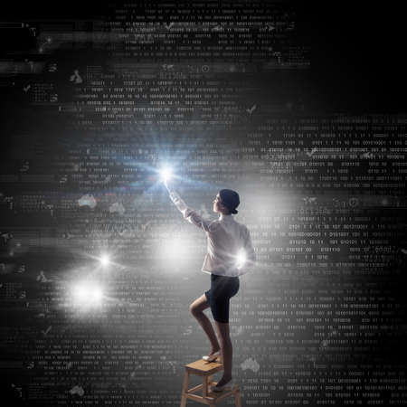 Back view of businesswoman standing on chair and reaching icon on virtual screenの写真素材