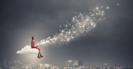 Young woman in dress with old book in hand sitting on cloudの写真素材