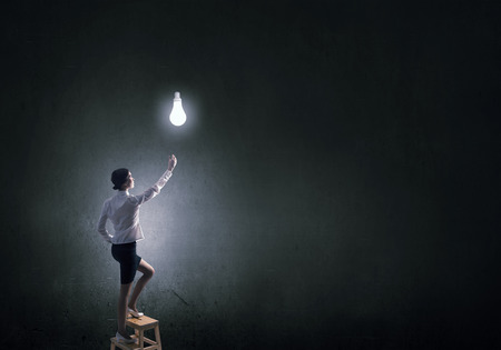 Back view of businesswoman standing on chair and reaching light bulbの写真素材