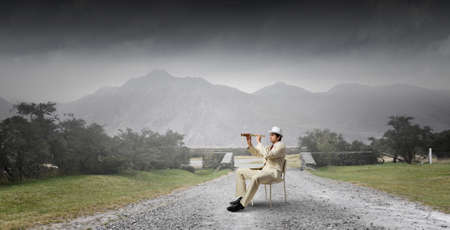 Businessman in white suit and hat sitting in chair and looking in spyglassの写真素材