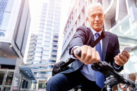 Successful businessman in suit riding bicycle and holding mobile phoneの写真素材